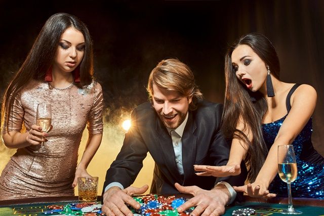 Two beautiful women and young man play on poker table in casino, focus on man and brunette